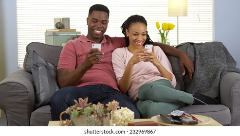 Happy Young Black Couple Relaxing On Couch Using Smartphones