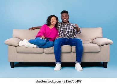 Happy Young Black Couple With Remote Control Sitting On Sofa And Watching TV Over Blue Studio Background, Full Length. African American Spouses With Controller Enjoying Television Program Or Show
