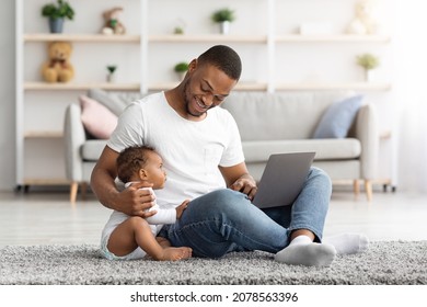 Happy Young Black Dad Using Laptop And Babysitting His Child At Home, Smiling African American Man Working Online Or Relaxing With Laptop In Living Room ANd Playing With Cute Infant Baby, Copy Space