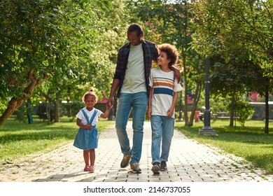 Happy Young Black Family Going On Pavement Road In Green Park. Father Hugging Son And Holding Hand Of Pleased Daughter. Relationship And Enjoy Time Together. Fatherhood And Parenting. Warm Sunny Day