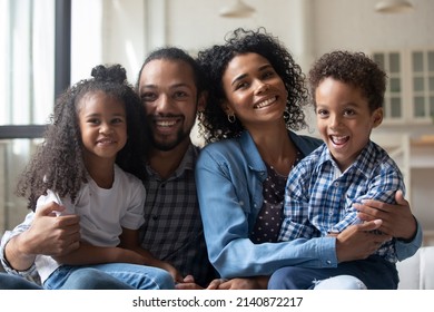 Happy Young Black Family Posing On Photo Shooting, Sitting On Couch, Looking Away, Smiling, Laughing. Millennial Couple Of Parents Holding Sibling Kids In Arms On Lap, Watching TV. Home Portrait