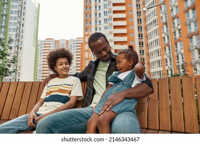 Happy Young Black Family Resting And Enjoying Time Together On Wooden Bench In Yard Of Modern District Outdoors. Father Hugging Little Daughter And Teenage Son. Fatherhood And Parenting. Relationship