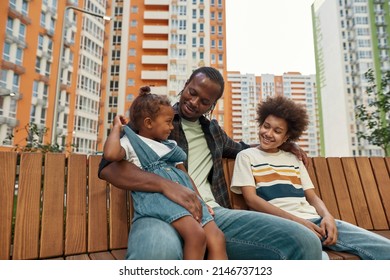 Happy Young Black Family Resting And Spending Time Together On Wooden Bench In Yard Outdoors. Father Hugging His Little Daughter And Teenage Son. Fatherhood And Parenting. Relationship. Warm Day