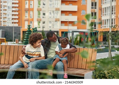 Happy Young Black Family Sitting And Resting On Wooden Bench In City Yard Outdoors. Father Hugging His Joyful Little Daughter And Son. Fatherhood And Parenting. Relationship And Spending Time Together