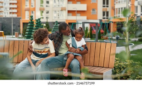 Happy Young Black Family Sitting And Resting On Wooden Bench In Yard Outdoors. Father Hugging His Pleased Daughter And Teenage Son. Fatherhood And Parenting. Relationship And Spending Time Together