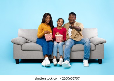 Happy Young Black Family Of Three People Watching Movie, Parents Sitting With Daughter On Couch, Eating Popcorn From Bucket Laughing At Show Holding Remote Control Isolated On Blue Studio Wall