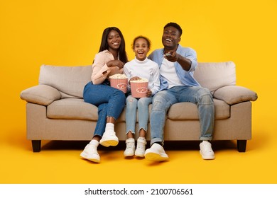 Happy Young Black Family Of Three People Watching Movie, Parents Sitting With Daughter On Sofa, Eating Popcorn From Bucket Laughing At Show Holding Remote Control Isolated On Yellow Orange Studio Wall