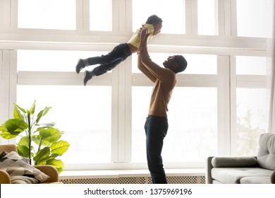 Happy young black father lifting up in air holding raise hands little son excited family playing active games having fun stands in living room near window people spending time together at home concept