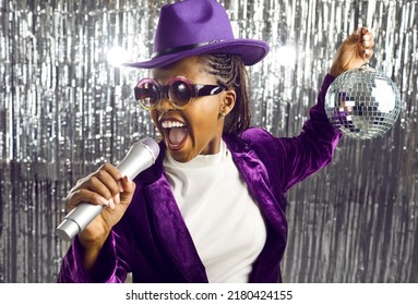 Happy young black girl having fun at karaoke party night. Cool excited funky African American woman singer in purple hat, jacket and party glasses holding silver disco ball and singing in microphone