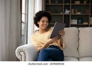 Happy young Black girl relaxing on sofa, using tablet computer, reading book, watching movie, media content, chatting online, talking on video call, laughing, smiling. Internet communication