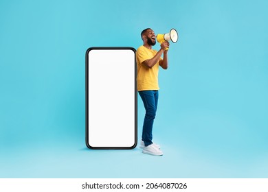 Happy young black guy shouting into megaphone, standing near big smartphone with empty screen, promoting your mobile app or website, offering mockup space for advertisement, blue background