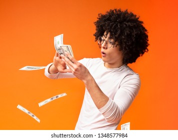 Happy young black guy throwing out money banknotes, orange studio background