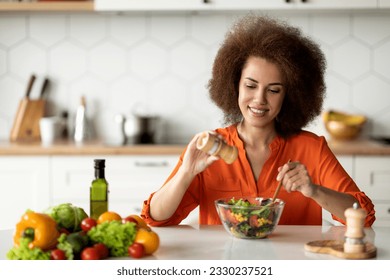 Happy young black lady preparing healthy vegetable salad in kitchen, seasoning meal, adding salt to bowl, smiling millenial african american female enjoying cooking food at home, free space - Shutterstock ID 2330237521