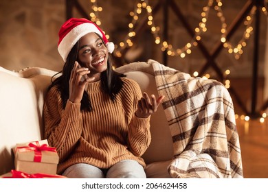 Happy young black lady in Santa hat sitting on couch, having mobile phone conversation on Xmas eve at home, free space. Millennial woman calling on smartphone, congratulating friends with Christmas - Powered by Shutterstock