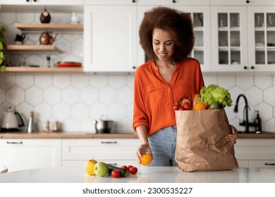 Happy Young Black Lady Unpacking Paper Bag With Groceries After Food Shopping, Smiling Millennial African American Woman Putting Fresh Vegetables And Fruits On Table, Standing In Kitchen At Home - Powered by Shutterstock