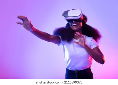 Happy young black lady in VR glasses exploring cyberspace, touching imaginary screen, playing virtual reality game in neon light. Positive African American woman experiencing contemporary technologies