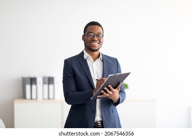 Happy young black male psychologist writing in clipboard, looking at camera and smiling in modern office. Psychotherapy services, mental health professional concept - Shutterstock ID 2067546356