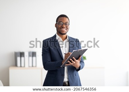 Happy young black male psychologist writing in clipboard, looking at camera and smiling in modern office. Psychotherapy services, mental health professional concept