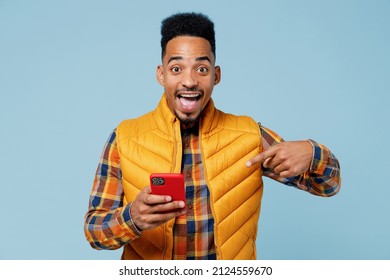 Happy young black man 20s years old wears yellow waistcoat shirthold in hand use mobile cell phone say wow just found out great win news isolated on plain pastel light blue background studio portrait