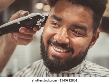 Happy Young Black Man Being Trimmed With Electric Clipper Machine In Barbershop.Male Beauty Treatment Concept.Smiling Young African Guy Getting New Haircut In Barber Salon.Portrait Of Barbers Client