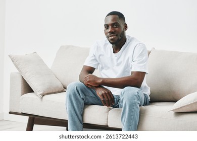 Hombre negro joven feliz en la camiseta blanca casual y los pantalones vaqueros que se sientan en un sofá moderno, sonriendo confiadamente en un ambiente luminoso, minimalista de la sala de estar
