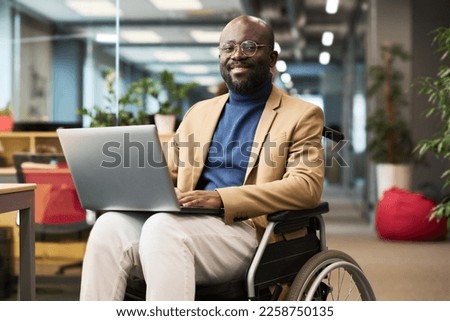 Happy young black man with disability sitting in wheelchair in office and looking at camera while analyzing online data on laptop screen