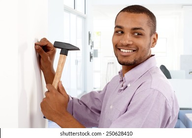 Happy Young Black Man Driving A Nail To Wall By Hammer, Smiling, Looking Away.