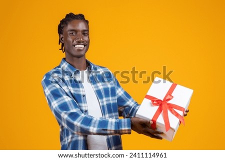 Happy young black man extending white gift box with red ribbon, cheerful african american guy offering present with friendly smile, standing against bright yellow background, free space