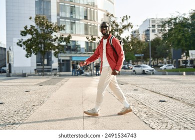 Happy young Black man in headphones listening music walking on city street. Cool stylish young African guy student hipster wearing sunglasses and backpack dancing outdoors. Candid photo. Full body. - Powered by Shutterstock