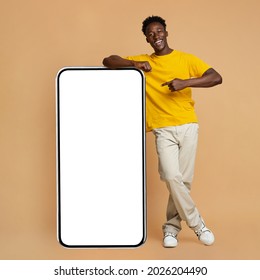 Happy Young Black Man Leaning And Pointing At Big Smartphone With White Blank Screen, Cheerful African Guy Showing Free Copy Space For Your Design, Standing Over Beige Background, Mockup Image