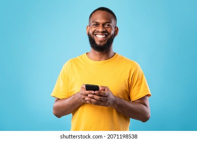 Happy Young Black Man Using Smartphone, Writing Message, Chatting With Friend, Surfing Internet, Watching Movie Online, Having Web Lesson Or Meeting On Blue Studio Background