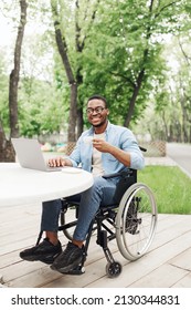 Happy Young Black Man In Wheelchair Using Laptop And Drinking Coffee At Table In Outdoor Cafe. Millennial African American Guy Working On Online Business Project At Park, Free Space