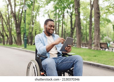 Happy Young Black Man In Wheelchair Using Tablet Pc, Browsing Web Or Watching Movie At Spring Park. Millennial African American Guy Checking Social Media On Mobile Device Outdoors