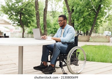 Happy Young Black Man In Wheelchair Using Laptop For Online Work At Cafe In Park, Having Business Conference On Web. African American Freelancer With Disability Working On Remote Project Outdoors