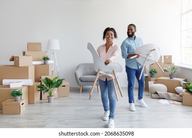 Happy Young Black Man And Woman Carry Chairs In New Home With Boxes. Couple Placing Furniture, Moving To New Flat, Family Planning House Improvement Interior, Design In Room Arrangement, Renovation