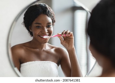 Happy Young Black Woman With Braces Covered In Towel Standing In Front Of Mirror In Bathroom, Brushing Her Teeth In The Morning, Closeup Shot. Dental, Oral, Teeth Care For Adults Concept