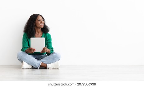 Happy Young Black Woman In Casual Sitting On Floor By White Wall With Modern Digital Tablet, Looking At Empty Space, Pretty African American Lady Surfing On Internet, Shopping Online, Panorama