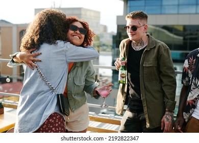 Happy young black woman with cocktail in martini glass giving hug to friend while standing against guy with bottle of beer - Powered by Shutterstock