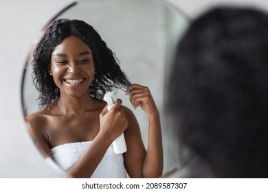 Happy young black woman covered in white towel using hair spray while styling her bushy hair after morning shower, looking at mirror and smiling, enjoying new hair care product, closeup