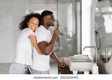 Happy Young Black Woman Hugging Her Husband From Back While Man Brushing His Teeth Near Mirror In Modern Bathroom Indoors, Enjoying Morning Beauty And Pampering Routine Together - Powered by Shutterstock