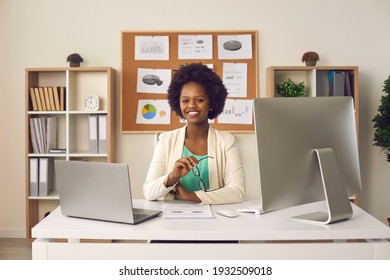 Happy young black woman looking at camera sitting at office table with laptop and desktop computer. Portrait of smiling businesswoman, head of company, business firm owner or economics teacher at work
