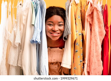 Happy Young Black Woman Peeking Out Between Clothes Hanging In Rail, Smiling At Camera, Satisfied With Shopping Choice, Buying Summertime Wardrobe Over Orange Studio Background