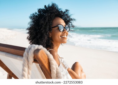 Happy young black woman relaxing on deck chair at beach wearing spectacles. Smiling african american girl with sunglasses enjoy vacation. Carefree happy young woman sunbathing at sea with copy space. - Shutterstock ID 2272553083