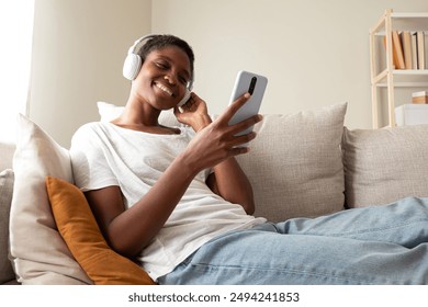 Happy young Black woman relaxing sitting on the couch using phone, listening to music with headphones. African American female at home texting with mobile phone. Lifestyle concept. - Powered by Shutterstock