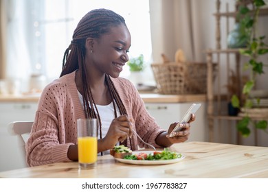 Happy Young Black Woman Scrolling Social Networks On Smartphone While Having Breakfast, Smiling African American Lady Eating Tasty Morning Meal And Browsing Internet On Mobile Phone, Free Space