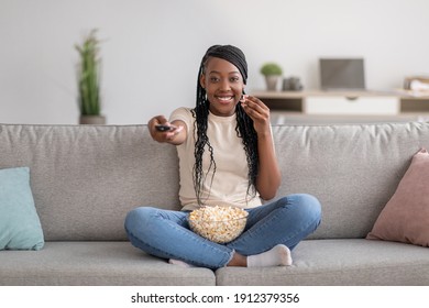 Happy young black woman sitting on couch with TV remote, choosing movie to watch and eating popcorn, copy space. Cheerful african american lady switching channels, home entertainment concept