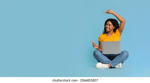 Happy Young Black Woman Sitting On Floor With Brand New Laptop, Pointing And Looking At Copy Space Over Blue Studio Background, Pretty African American Lady Using Notebook, Panorama