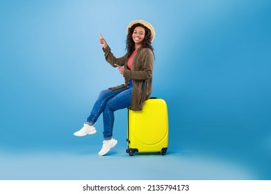 Happy young black woman sitting on bright suitcase and pointing at empty space for travel agency ad on blue studio background. Cheerful female traveler with luggage going on summer vacation