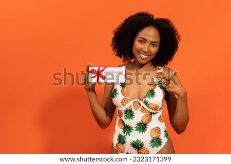 Happy young black woman slim body wear swimsuit hold gift certificate coupon voucher store card isolated on orange color background studio, copy space. Summer hotel pool sea rest sun tan concept