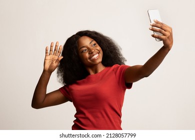 Happy Young Black Woman With Smartphone Making Video Call Or Taking Selfie, Portrait Of Cheerful African American Female Waving Hand At Mobile Phone Camera While Standing Over White Studio Background - Powered by Shutterstock